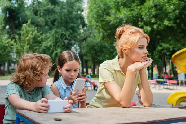 Mujer pensativa y triste mirando hacia otro lado cerca de los niños jugando con los teléfonos inteligentes en el parque - foto de stock