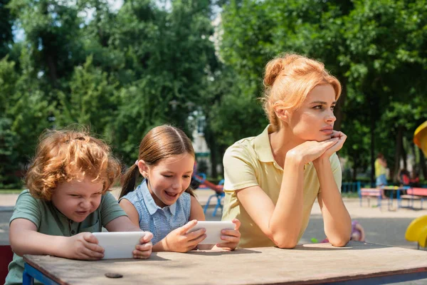 Niños emocionados jugando en teléfonos móviles cerca de madre molesta al aire libre - foto de stock