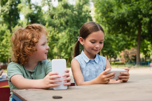 Chico y chica positiva usando teléfono inteligente mientras está sentado en el parque - foto de stock