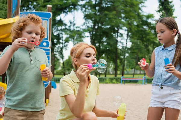 Frau pustet mit Kindern Seifenblasen auf Spielplatz — Stockfoto