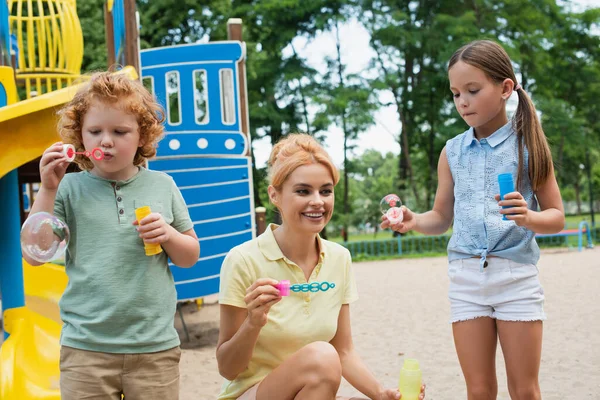 Femme joyeuse avec ventilateur à bulles près des enfants sur l'aire de jeux — Photo de stock