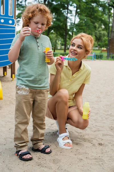 Full length view of happy woman near son blowing soap bubble on playground — Stock Photo