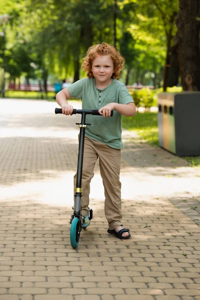 Vue complète de l'enfant heureux avec trottinette de coup de pied regardant la caméra dans le parc vert — Photo de stock