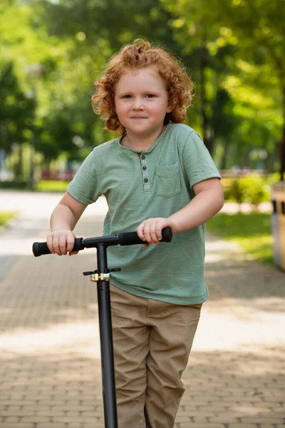 Menino encaracolado com chute scooter sorrindo para a câmera ao ar livre — Fotografia de Stock