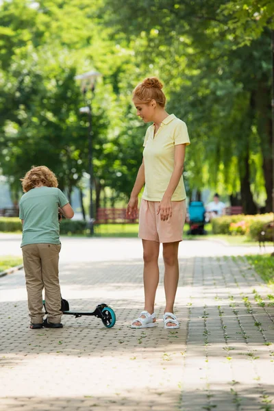 Pleine longueur vue de la femme en vêtements d'été debout près de fils avec kick scooter dans le parc — Photo de stock