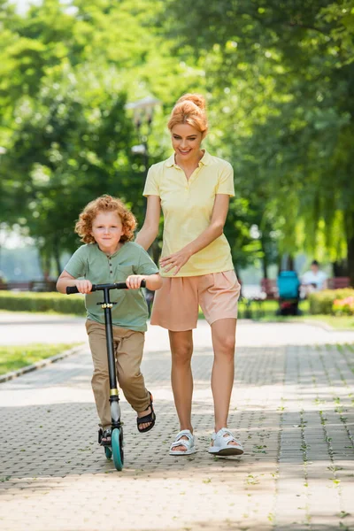 Volle Länge Ansicht der Rotschopf Junge Reiten Kick Roller in der Nähe Mama in Park — Stockfoto