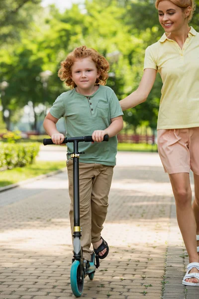 Rothaariger Junge schaut in die Kamera, während er Tretroller in der Nähe lächelnder Mutter fährt — Stockfoto