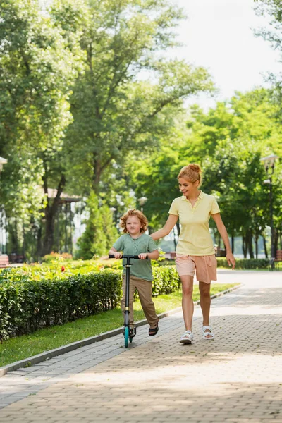 Visão de comprimento total da mulher em roupas de verão andando perto do filho montando chute scooter no parque — Fotografia de Stock