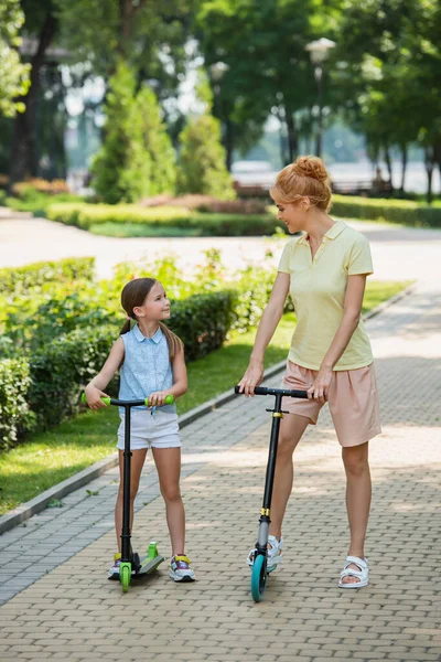 Visão de comprimento total de mãe e filha olhando um para o outro enquanto montando scooters chute no parque — Fotografia de Stock