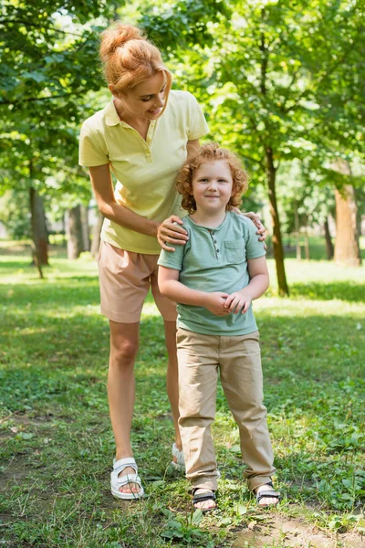 Full length view of woman embracing shoulders of son while standing on lawn in park — Stock Photo