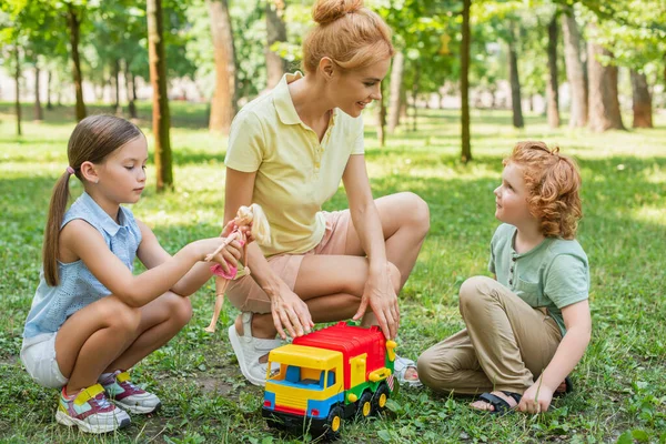 Enfants jouer avec poupée et jouet camion près de sourire maman sur la pelouse verte — Photo de stock