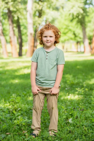 Позитивный мальчик в грязных брюках стоит на газоне и смотрит в камеру — стоковое фото