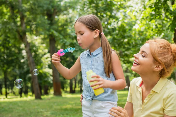 Donna allegra sorridente vicino a figlia che soffia bolle di sapone nel parco — Foto stock