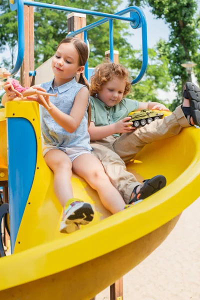 Vue complète du frère et de la sœur assis sur la glissière et jouant avec des jouets — Photo de stock