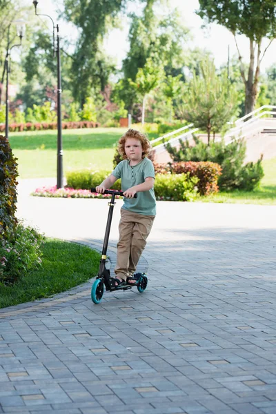 Full length view of curly boy riding kick scooter in summer park — Stock Photo