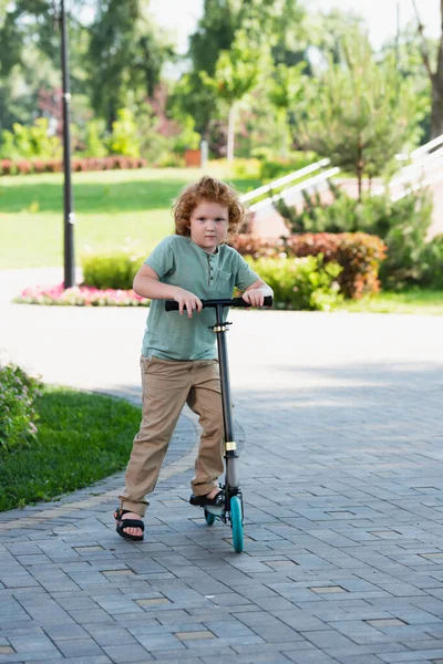 Pleine longueur vue de rousse garçon avec coup de pied scooter regardant caméra dans le parc — Photo de stock