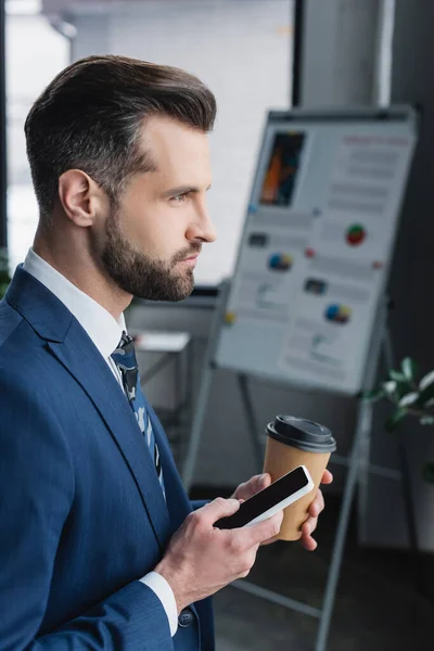 Vue latérale de l'économiste tenant tasse en papier et smartphone avec écran blanc dans le bureau — Photo de stock