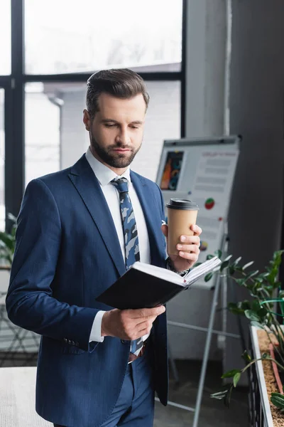 Economista morena con café para ir leyendo cuaderno en la oficina - foto de stock