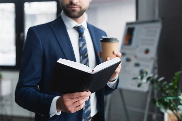 Vista ritagliata di uomo d'affari offuscata con notebook e tazza di carta in ufficio — Foto stock