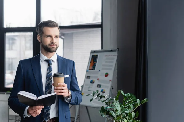 Hombre de negocios con bebida para llevar y cuaderno mirando hacia otro lado en la oficina - foto de stock