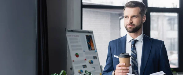 Экономист с кофе, чтобы уйти, глядя в сторону размытого флип-графика в офисе, баннер — стоковое фото