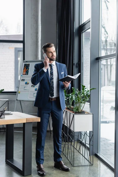 Vue complète de l'économiste avec ordinateur portable parlant sur le téléphone mobile dans le bureau — Photo de stock