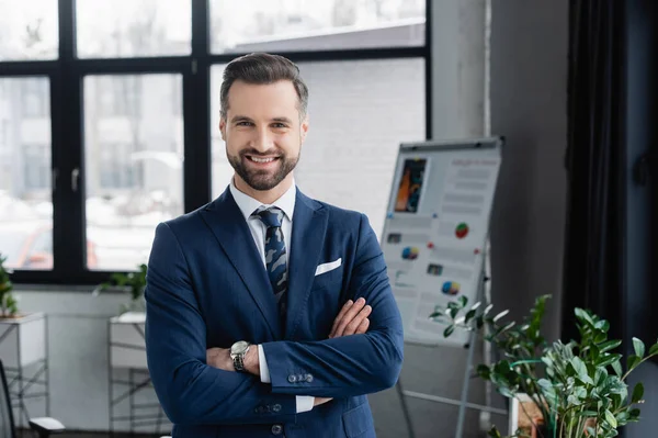 Glückliche brünette Ökonomin im Blazer mit verschränkten Armen stehend und lächelnd in die Kamera — Stockfoto