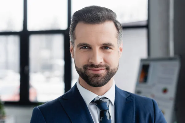 Barbuto uomo d'affari sorridente a macchina fotografica in ufficio offuscata — Foto stock