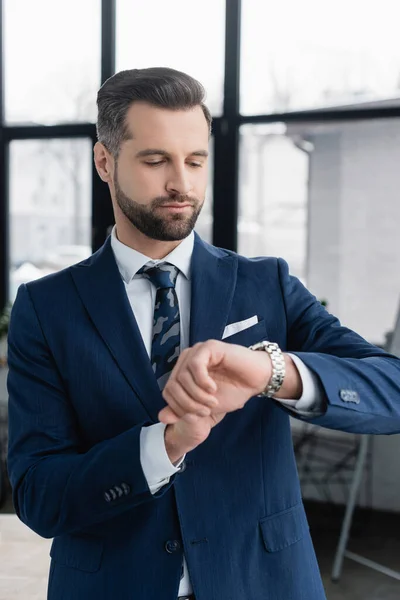 Hombre de negocios en blazer mirando reloj de pulsera en oficina borrosa — Stock Photo