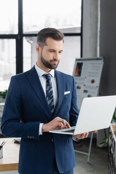 Economist in suit using laptop while standing in blurred office — стоковое фото