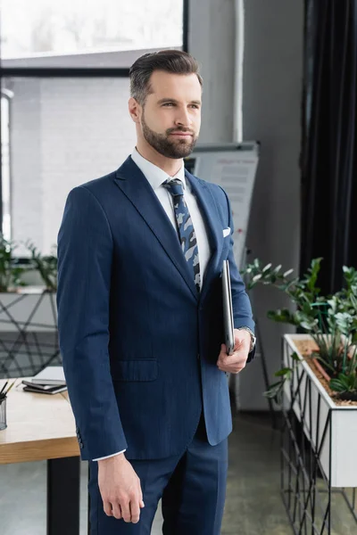 Der bärtige Ökonom schaut weg, während er mit Laptop im Büro steht — Stockfoto