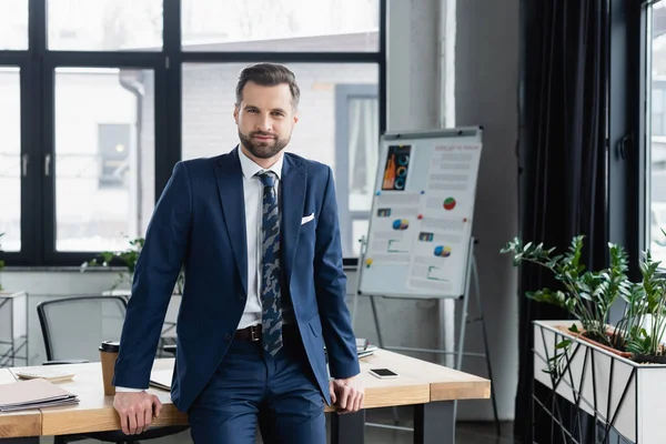 Positiver Ökonom steht im Büro am Schreibtisch und blickt in die Kamera — Stockfoto