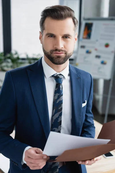 Brunette businessman with documents looking at camera in blurred office — Stock Photo