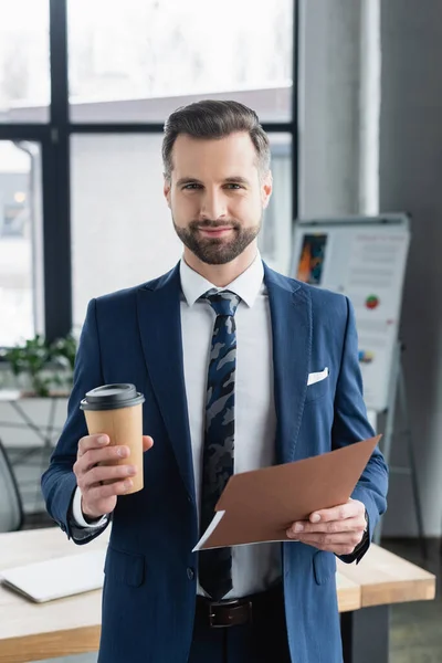 Lächelnder Ökonom mit Coffee to go und Aktenordner, der im modernen Büro in die Kamera blickt — Stockfoto