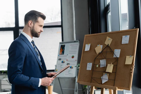 Side view of economist with folder near corkboard with sticky notes and blurred flip chart — стоковое фото