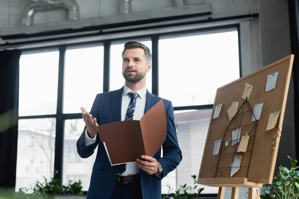 Economista con carpeta apuntando con la mano y hablando cerca de corcho con notas adhesivas — Stock Photo