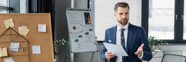 Ökonom zeigt mit der Hand, während er mit Papier in der Nähe von Korkplatten steht und Grafiken auf Flipchart, Banner — Stockfoto