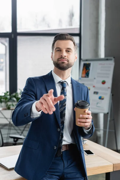 Ökonom mit Pappbecher blickt in Kamera und zeigt mit Finger in verschwommenes Büro — Stockfoto