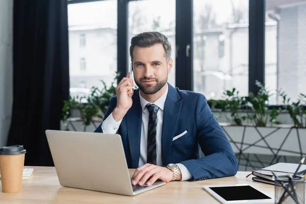 Hombre de negocios positivo mirando a la cámara mientras habla en el teléfono inteligente cerca de la computadora portátil - foto de stock