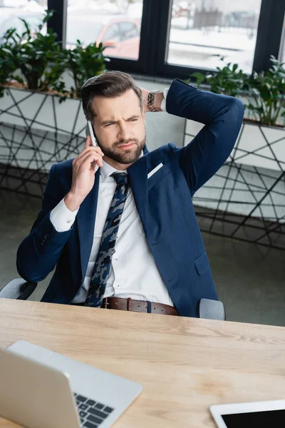 Pensativo hombre de negocios mirando hacia otro lado mientras habla en el teléfono inteligente en la oficina - foto de stock