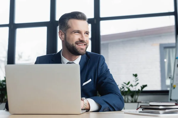 Economista alegre mirando hacia otro lado mientras trabaja cerca de la computadora portátil y portátiles - foto de stock