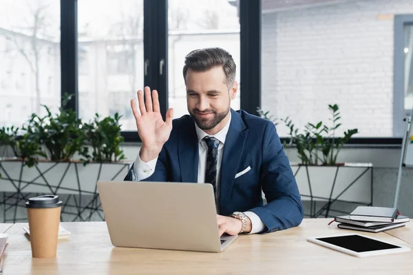 Economista sonriente saludando la mano durante la videollamada en el ordenador portátil en la oficina — Stock Photo