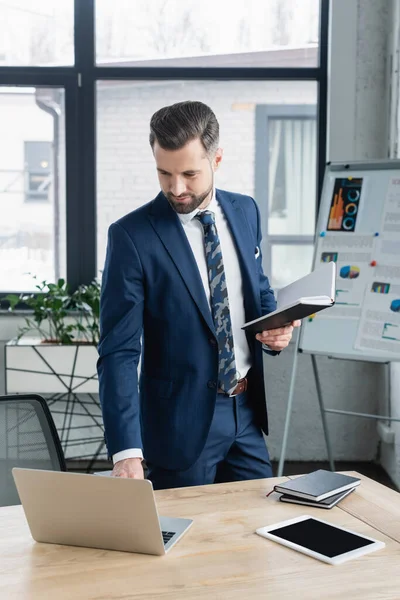 Economista que utiliza el ordenador portátil mientras está de pie con portátil en el lugar de trabajo - foto de stock