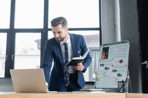 Economista con notebook guardando computer portatile vicino flip chart su sfondo sfocato — Foto stock