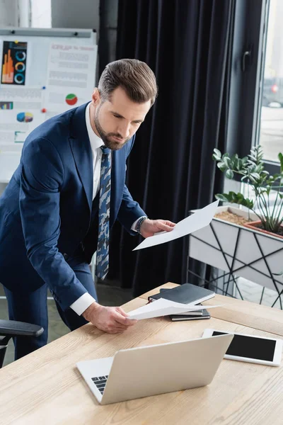 Businessman in suit holding papers and looking at laptop in office — Stock Photo