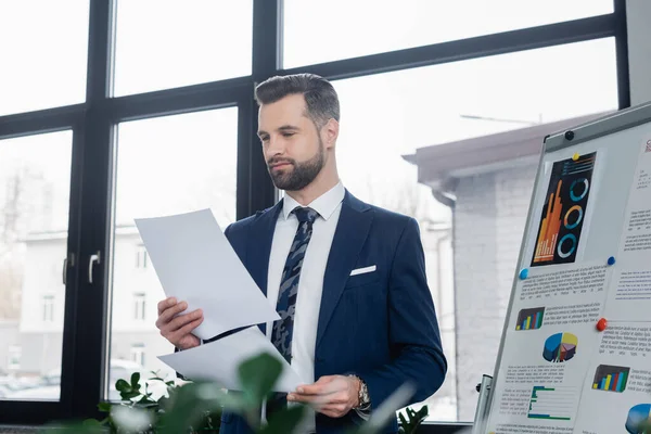 Economista barbudo olhando para papéis perto de flip chart com gráficos no escritório — Fotografia de Stock