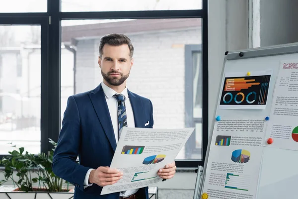 Bearded economist with analytics looking at camera near flip chart in office — Stock Photo