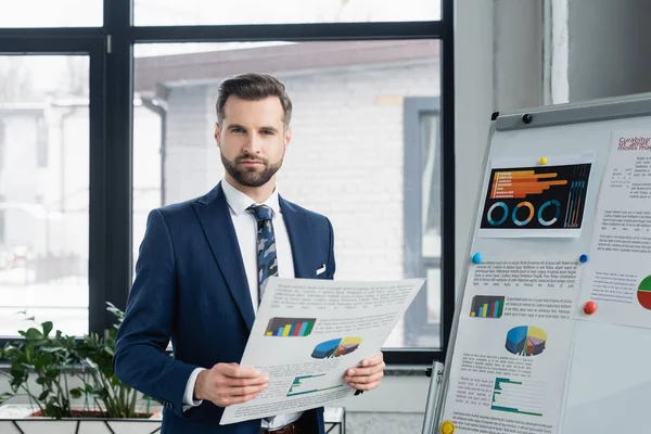 Bearded economist with analytics looking at camera near flip chart in office — стоковое фото