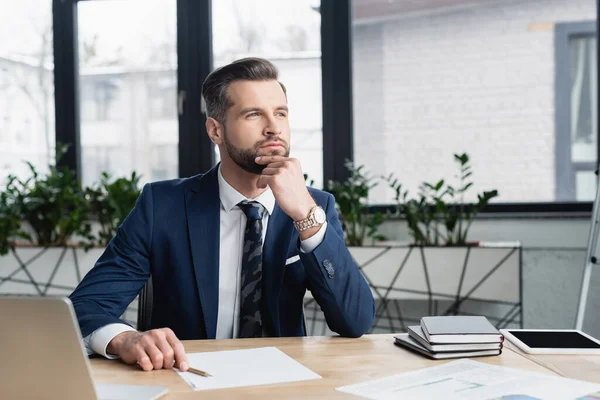 Nachdenklicher Ökonom schaut weg, während er neben Dokumenten und Notizbüchern sitzt — Stockfoto