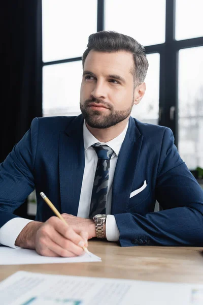 Thoughtful economist in blazer and tie looking away while writing in office — Stock Photo